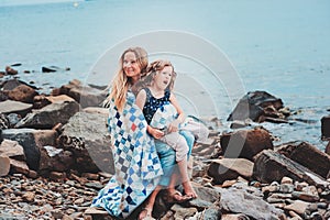 Happy mother and daughter wrapped in quilt blanket spending time together on the beach on summer vacation. Happy family traveling