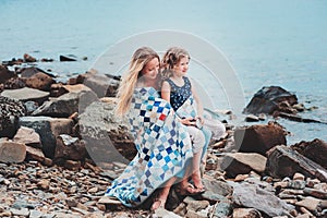 Happy mother and daughter wrapped in quilt blanket spending time together on the beach on summer vacation. Happy family traveling