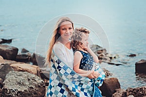 Happy mother and daughter wrapped in quilt blanket spending time together on the beach on summer vacation. Happy family traveling