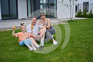 Happy mother, father and daughter sitting on grass and looking at camera in yard