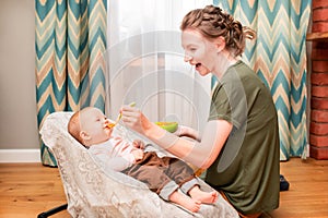 Happy mother feeds laughing baby vegetable puree from plastic spoon at home