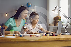 Happy mother helping her cute daughter study online at home. Parent and child watching art lesson together on laptop photo