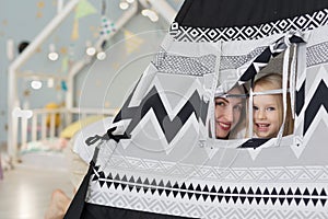 Happy mother with her 3 years old girl watching out of tent playhouse in playroom