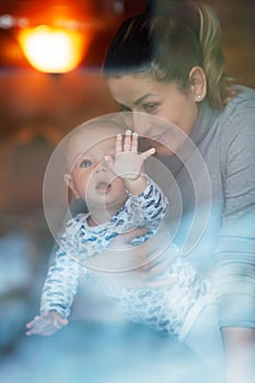 Happy mother and her baby boy looking throw the window and playing in warm home on winter day. Christmas time