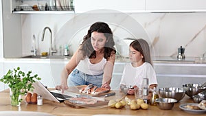 Happy mother and her daughter preparing salmon in the kitchen while looking at the recipe on their laptop