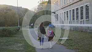 Happy mother with her daughter in school uniform go home. Mom and daughter are walking along the alley along the school