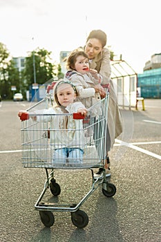 Happy mother and her daughters are having fun with a shopping cart on a parking lot beside a supermarket.