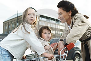 Happy mother and her daughters are having fun with a shopping cart on a parking lot beside a supermarket.