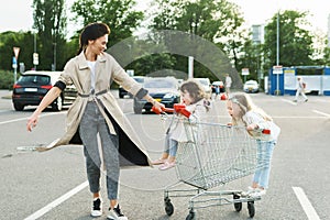 Happy mother and her daughters are having fun with a shopping cart on a parking lot beside a supermarket.