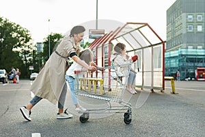 Happy mother and her daughters are having fun with a shopping cart on a parking lot beside a supermarket.