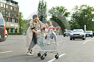 Happy mother and her daughters are having fun with a shopping cart on a parking lot beside a supermarket.