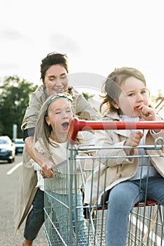 Happy mother and her daughters are having fun with a shopping cart on a parking lot beside a supermarket.