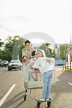 Happy mother and her daughters are having fun with a shopping cart on a parking lot beside a supermarket.