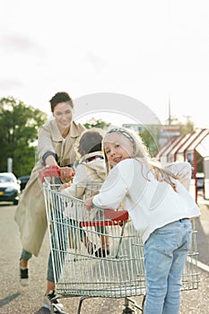 Happy mother and her daughters are having fun with a shopping cart on a parking lot beside a supermarket.