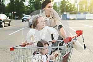 Happy mother and her daughters are having fun with a shopping cart on a parking lot beside a supermarket.