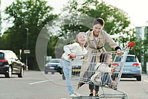 Happy mother and her daughters are having fun with a shopping cart on a parking lot beside a supermarket.