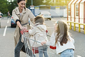 Happy mother and her daughters are having fun with a shopping cart on a parking lot beside a supermarket.