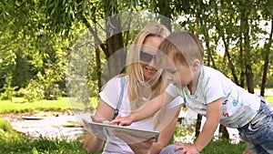 Happy mother and her little son enjoying playing on tablet computer in the park. Slow motion.