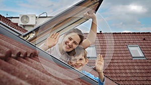 Happy mother and her son open attic windows, embrace the view outside, and warmly wave towards the camera. Family