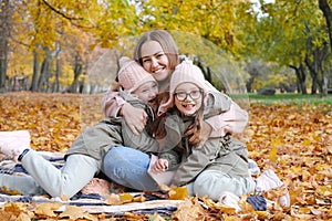 A happy mother with her twin daughters spends time sitting on a blanket in an autumn park. Mother hugging her daughters