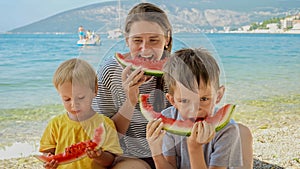 Happy mother and her two boys share laughter and sweet watermelon on a beachside picnic. Family vacation, the happiness
