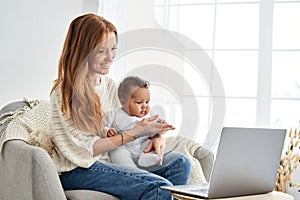 Happy mother holding cute baby daughter looking at laptop computer at home.