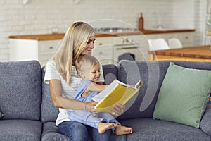 Happy mother holding her baby boy on knees and reading book together at home on sofa