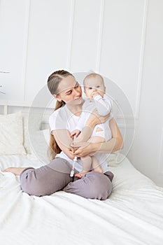 Happy mother holds baby in her arms playing with him and hugging him on the bed at home