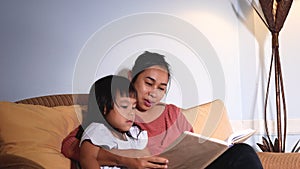 Happy mother hugging cute little daughters reading a book fairy tale together sitting on couch at home. Bedtime Story Concept.