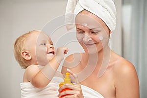 Happy mother and infant baby in white towels after bathing apply sunscreen or after sun lotion. Children skin care in a hotel or