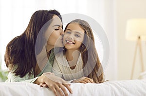 Happy mother kisses her pretty little daughter while cuddling together on a bed at home