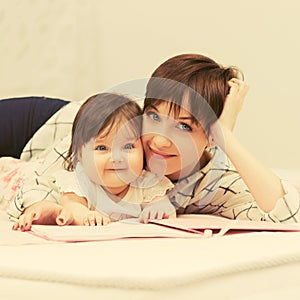 Happy mother and little baby girl reading a book on bed