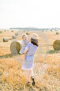 Happy mother and little baby girl standing next to hay bales in harvested field in sunny summer day. Mother and daughter