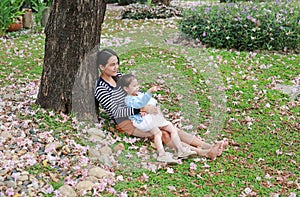 Happy mother and little child girl lying on green grass with fall pink flower in the garden outdoor. Asian girl pointing for her