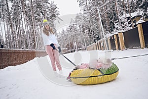 Happy mother and little cute girl in pink warm outwear walking having fun rides inflatable snow tube in snowy white