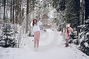 Happy mother and little cute girl in pink warm outwear walking playing snowball fight having fun in snowy white winter coniferous