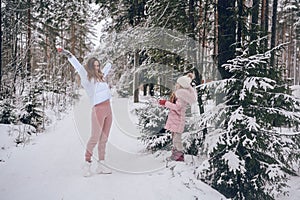 Happy mother and little cute girl in pink warm outwear walking playing snowball fight having fun in snowy white winter coniferous