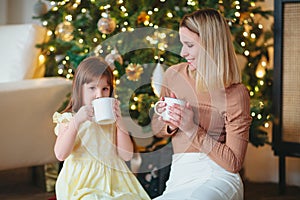 Happy mother and little daughter drinking cacao while sitting near decorated ?hristmas tree at home