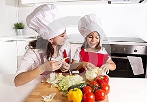 Happy mother and little daughter at home kitchen preparing salad in apron and cook hat