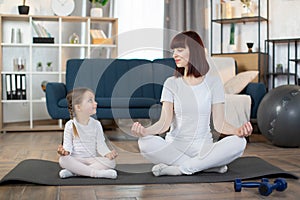 Happy mother with little daughter meditating, sitting in easy seat pose on mat at home