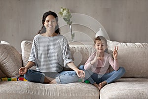 Happy mother and little daughter practicing yoga, meditating on couch
