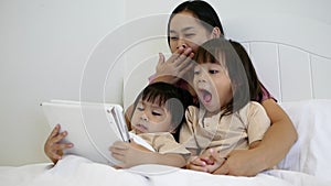 Happy mother and little daughters playing tablet pc computer on bed at night. Happy family time.