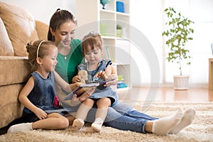 Happy mother and little daughters reading a book together in the living room at home. Family activity concept.
