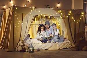 Happy mother and little daughters sitting in cozy playroom tent and reading book together