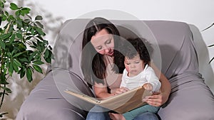 Happy mother and Mixed-race toddler little son reading book on couch at home.