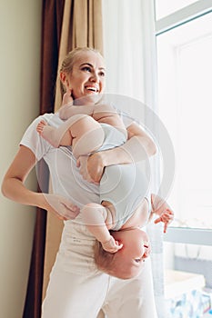 Happy mother playing with her baby son at home holding infant upside down. Family having fun