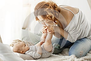 Happy mother playing with newborn baby kissing little legs spending best maternity moments in cozy bedroom. Warm family