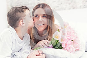 Happy mother`s day concept. Mom with son on bed with gift and tulips.Son kisses his mom