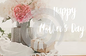 Happy mother`s day lettering on pink and white peonies in vase and gift box with on rustic table in light, space for text. Happy