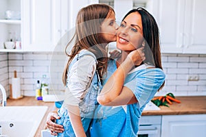 Happy Mother& x27;s Day. Little daughter kissing and hugging her smiling mom in light modern kitchen together